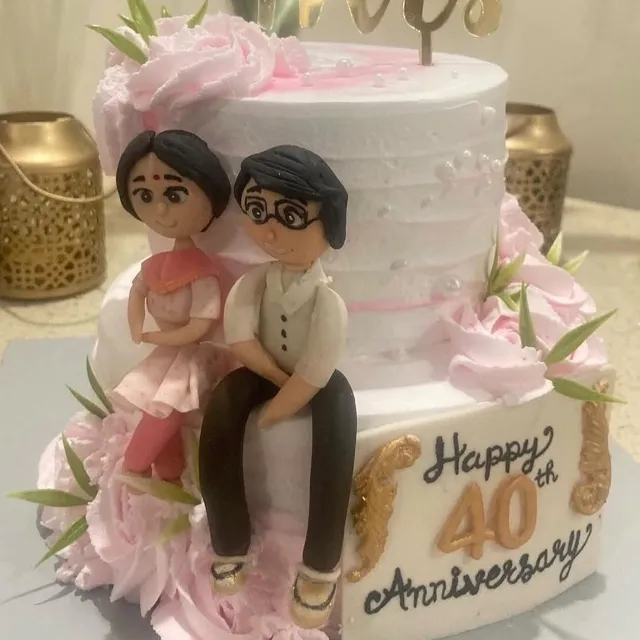 A 40th Anniversary Cake — A two-tier white cake with pink buttercream ruffle roses, a gold “Mrs” topper, hand-sculpted figures of the couple, and gold-lettered “Happy 40th Anniversary.”