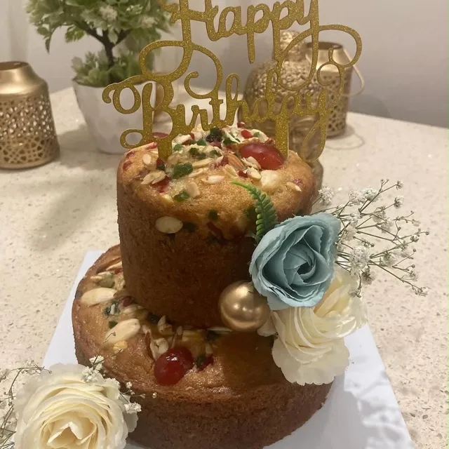 A Two-Tier Dry Cake for Puneet — A two-tier fruit dry cake topped with almonds, glacé cherries, and candied peel, finished with fresh blue and cream roses, a gilded ball, and a gold glitter topper.
