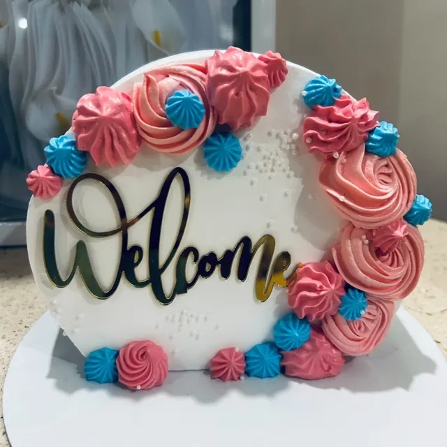 A Welcome Rosette Cake — A hand-piped rosette wreath in blush and sky blue, finished with a mirrored gold Welcome topper.